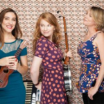 Moria Smiley and two other women musicians standing in front of a patterned wallpaper.